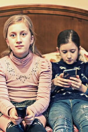 Little girls with smartphone watch TV. The girl in front of the TV looking in the smartphone.