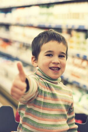 Happy boy in a supermarket on the background of shelves with food. The boy showed that everything is ok. toned. vertical photoの写真素材