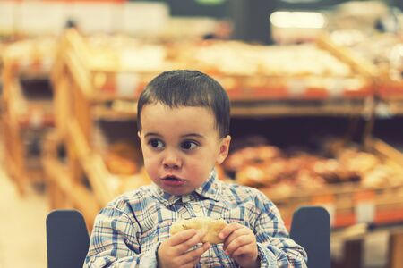 Cute boy eating a bun or pita bread in a supermarket in the cart. Hungry child in the store. tonedの写真素材