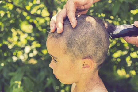 hand holds an electric hair clipper and cuts the boy's long hair, barbershop at home, parent cuts...