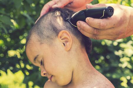 hand holds an electric hair clipper and cuts the boy's long hair, barbershop at home, parent cuts hair while hairdressers are closed, stay home concept. toned.の写真素材