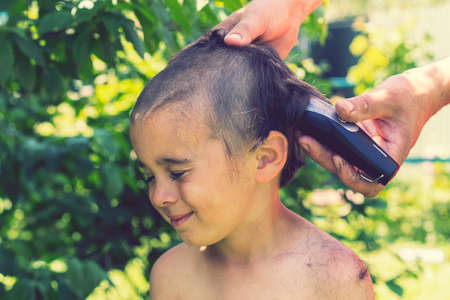 hand holds an electric hair clipper and cuts the boys long hair, barbershop at home, parent cuts hair while hairdressers are closed, stay home concept. tonedの写真素材