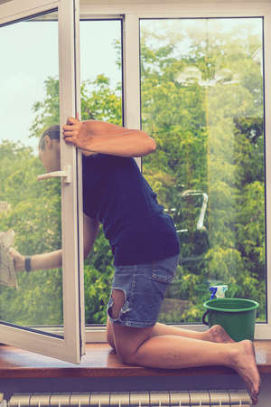 Back view of the young woman who washing the window with rag and window cleaner in the room. Cute girl with long hair washes a glass with detergent. toned. selected focus. tonedの写真素材