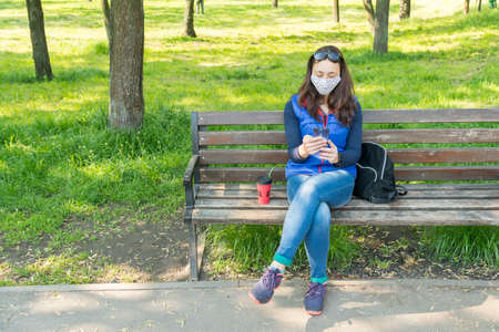 The woman sitting on the bench in the park and wearing a protective mask and using smartphone .の写真素材