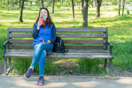 Beautiful Caucasian woman with long brown hair sitting on a yoga mat in the park drinking a cup of tea or coffee and smiling. Middle-aged athletic woman with long hair drinks coffee in a parkの写真素材