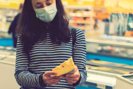 woman in protective medical mask in a supermarket chooses cheese. pandemic concept. Hand of the buyer with a piece of cheese in the store. tonedの写真素材
