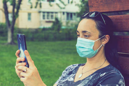 Portrait of a Girl with medical mask at park with smartphone during quarantine. A young girl in a medical mask sits on bench in the park and use smartphone. tonedの写真素材