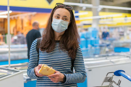woman in protective medical mask in a supermarket chooses cheese. pandemic and covid-19 concept. Hand of the buyer with a piece of cheese in the store. tonedの写真素材