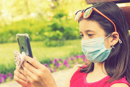 girl in protective mask holding a smartphone. teen girl siting on bench in green park with mobile phone. First stage of loosening coronavirus restrictions and self-isolation. tonedの写真素材