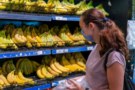 Focused woman in face mask choosing fruits, taking bananas from shelves in grocery store. Customer in supermarket. Side view. Shopping during epidemic concept. tonedの写真素材