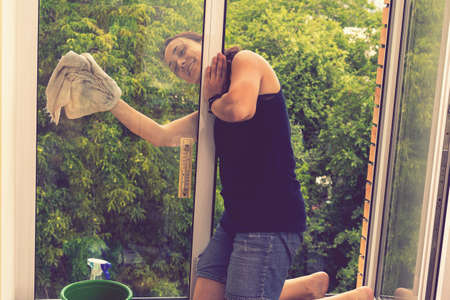 Back view of the young woman who washing the window with rag and window cleaner in the room. Cute girl with long hair washes a glass with detergent. toned. selected focus. tonedの写真素材