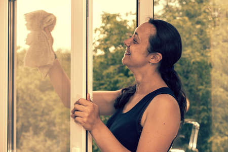 Back view of the young woman who washing the window with rag and window cleaner in the room. Cute girl with long hair washes a glass with detergent. toned. selected focus. tonedの写真素材