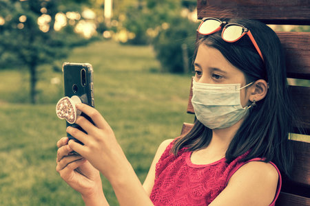 girl in protective mask holding a smartphone. teen girl siting on bench in green park with mobile phone. First stage of loosening coronavirus restrictions and self-isolation. tonedの写真素材