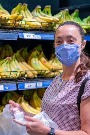 Focused woman in face mask choosing fruits, taking bananas from shelves in grocery store. Customer in supermarket. Side view. Shopping during epidemic concept. vertical photoの写真素材