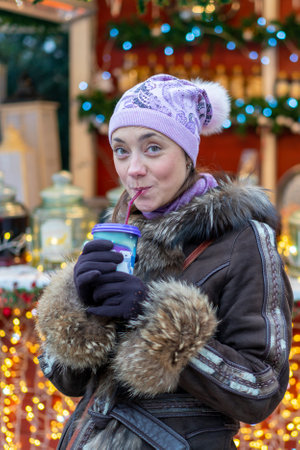 Beautiful young woman with coffee cup in the city. Beautiful woman with coffee on the street in winter clothes. Woman Having Hot Drink Outdoors On Winter Market. toned. vertical photoの写真素材