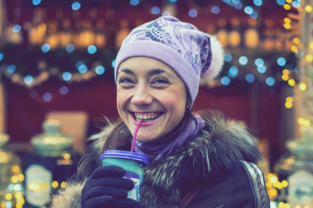 Beautiful young woman with coffee cup in the city. Beautiful woman with coffee on the street in winter clothes. Woman Having Hot Drink Outdoors On Winter Market. tonedの写真素材