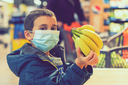 Child wearing surgical face mask buying fruit in supermarket in pandemic. little boy in a supermarket is wearing a medical mask. quarantine. toned.の写真素材