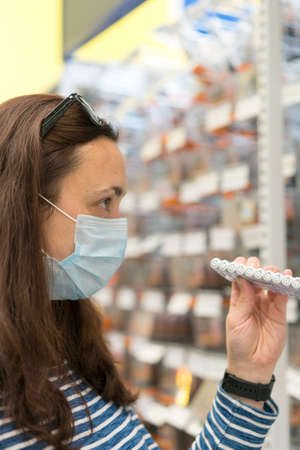 Woman in medical mask choosing goods in a store. Concept of shopping during quarantine at virus pandemic. shopping at the time of the virus. Beautiful young women in a flu maskの写真素材