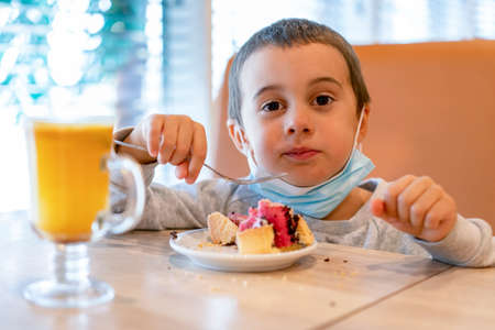 Little boy in a medical mask in a cafe eats a cake. A child wearing a mask in a cafe during a...
