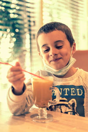 A child wearing a mask in a cafe during a pandemic. A boy in a cafe drinks sea buckthorn tea in a cafe during a pandemic. toned. vertical photoの写真素材