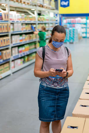 Woman wearing face mask and shopping in grocery store. Concept of protection against virus and communication. Adult woman in medical mask using smartphone and shopping for groceriesの写真素材