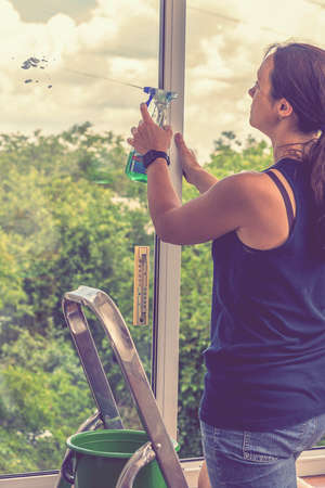 Attractive Woman Washing the Window. Cleaning Company worker working. Young woman washing window, close up. Cute girl with collected hair washes a glass with detergent. selected focus. tonedの写真素材