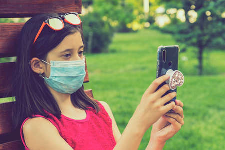girl in protective mask holding a smartphone. teen girl siting on bench in green park with mobile phone. First stage of loosening pandemic restrictions and self-isolation. tonedの写真素材