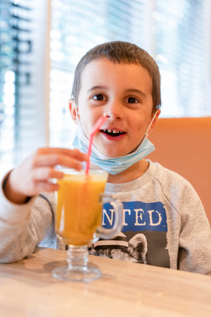 A child wearing a mask in a cafe during a pandemic. A boy in a cafe drinks sea buckthorn tea in a cafe during a pandemicの写真素材