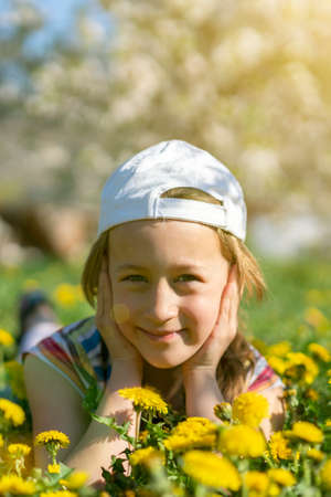 Closeup portrait of little cute girl lying with dandelions on the grass. Happy smiling girl lies on the grass among yellow flowering dandelions in the park. toned. vertical photo.の写真素材
