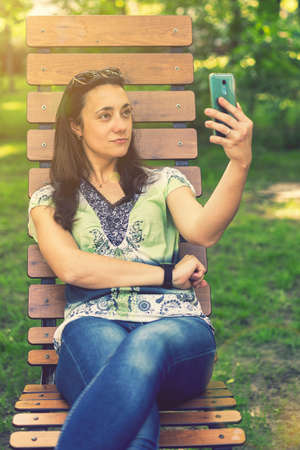 Cheerful girl after shopping with many shopping bags sitting on bench in the park and taking selfie with smart phone. Young woman takes a selfie, sits on a bench in a city park. toned. vertical photoの写真素材