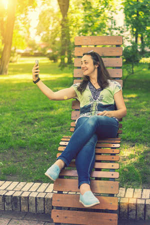 Young woman takes a selfie, sits on a bench in a city park. toned. vertical photoの写真素材