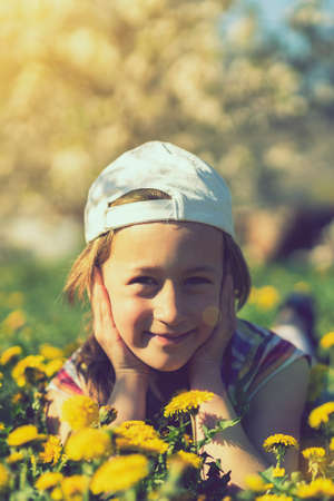 Closeup portrait of little cute girl lying with dandelions on the grass. Happy smiling girl lies on the grass among yellow flowering dandelions in the park. toned. vertical photoの写真素材
