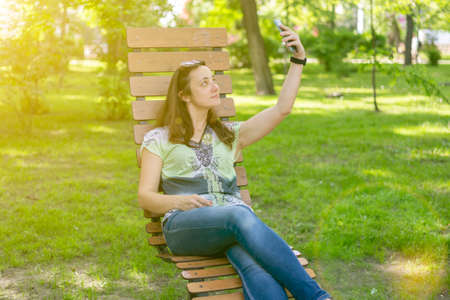 Cheerful girl after shopping with many shopping bags sitting on bench in the park and taking selfie with smart phone. Young woman takes a selfie, sits on a bench in a city park. tonedの写真素材