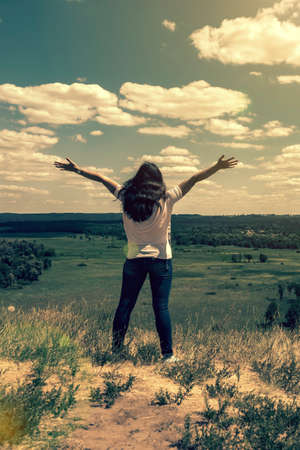 Young woman standing in a wheat field with sunrise on the background. A view from the back of a women standing on a mountain and admires the beautiful view sunset. toned. vertical photoの写真素材