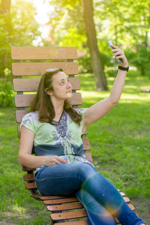 Young woman takes a selfie, sits on a bench in a city park. toned. vertical photoの写真素材