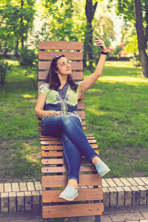 Cheerful girl after shopping with many shopping bags sitting on bench in the park and taking selfie with smart phone. Young woman takes a selfie, sits on a bench in a city park. toned. vertical photoの写真素材