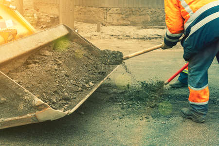 Road workers in bright orange reflective uniforms use shovels to scrape accumulated sand. maintenance of road and highway pavements. pothole repairing works. tonedの写真素材