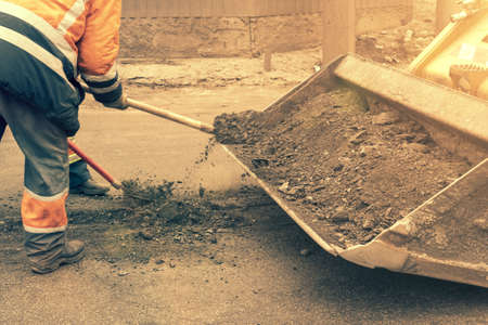Traffic workers in bright orange reflective uniforms scrape off accumulated sand and debris between lanes with shovels and load it into a metal grader bucket. toned.の写真素材