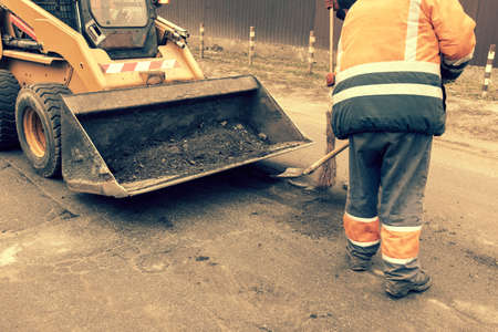 Road workers in bright orange reflective uniforms use shovels to scrape accumulated sand. maintenance of road and highway pavements. pothole repairing works. toned.の写真素材