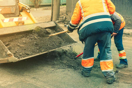 Road workers in bright orange reflective uniforms use shovels to scrape accumulated sand. maintenance of road and highway pavements. pothole repairing works. tonedの写真素材