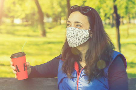 Woman relaxing in a spring park in a protective surgical face mask sitting on a bench in the sunshine during the coronavirus drinking coffee. tonedの写真素材