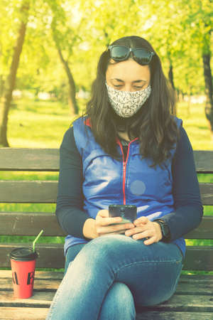 Portrait of brunette woman in a protective mask sitting on a bench in the park, sunny spring day. oronavirus concept. respiratory protection. vertical photo. tonedの写真素材