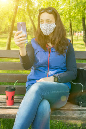 Portrait of brunette woman in a protective mask sitting on a bench in the park, sunny spring day. oronavirus concept. respiratory protection. vertical photo. tonedの写真素材