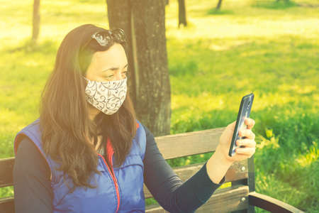 The woman sitting on the bench in the park and wearing a protective mask and using smartphone .の写真素材