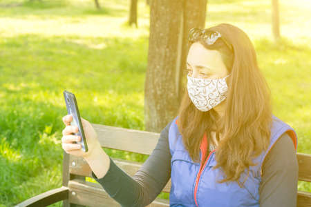 The woman sitting on the bench in the park and wearing a protective mask and using smartphone .の写真素材