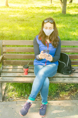 The woman sitting on the bench in the park and wearing a protective mask and using smartphone .の写真素材