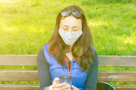 The woman sitting on the bench in the park and wearing a protective mask and using smartphone .の写真素材