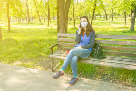 The woman sitting on the bench in the park and wearing a protective mask and using smartphone .の写真素材