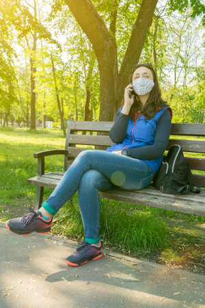 The woman sitting on the bench in the park and wearing a protective mask and using smartphone .の写真素材