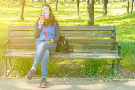 Beautiful Caucasian woman with long brown hair sitting in the park wearing a protective mask drinking a cup of tea or coffee and smiling. tonedの写真素材